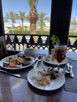 A dining table set with plates of assorted food including salads, noodles, grilled meat, and vegetables. There is a glass of iced tea next to the plates. Through the window, palm trees and a clear blue sky are visible, suggesting a tropical or Mediterranean atmosphere.