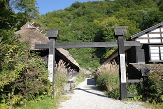 A tranquil rural setting features traditional thatched-roof houses lined along a gravel path. A large wooden gate stands at the entrance, accompanied by lush green vegetation and tall grasses on both sides. In the background, a dense forest covers a hill, with blue sky peeking through the foliage.