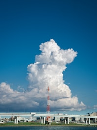 A tall communication tower stands in front of a backdrop of large, fluffy white clouds. The scene is under a clear blue sky, with a bridge and cityscape in the distance. The water in the foreground reflects the structures and sky.