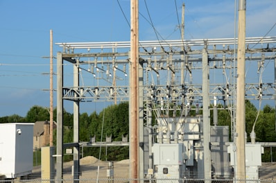 An electrical substation with large transformers and metal structures surrounded by a chain-link fence. Power lines and poles are visible against a clear blue sky. In the background, there are trees and a few residential homes.