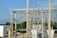 An electrical substation with large transformers and metal structures surrounded by a chain-link fence. Power lines and poles are visible against a clear blue sky. In the background, there are trees and a few residential homes.