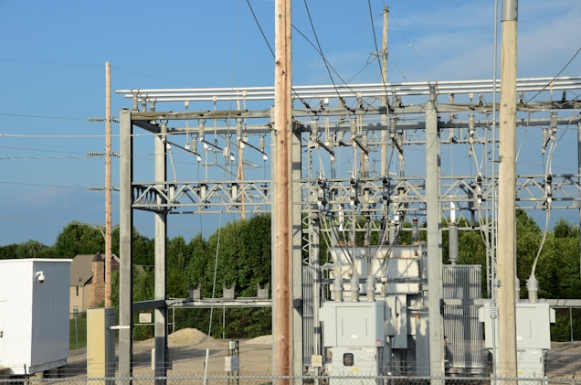 An electrical substation with large transformers and metal structures surrounded by a chain-link fence. Power lines and poles are visible against a clear blue sky. In the background, there are trees and a few residential homes.