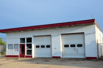 A small garage building with two large closed roller doors and a red door. The structure has a red trim on the roof, and the walls appear to be painted white. The ground is paved, and there is minimal vegetation visible near the garage.