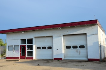 A small garage building with two large closed roller doors and a red door. The structure has a red trim on the roof, and the walls appear to be painted white. The ground is paved, and there is minimal vegetation visible near the garage.