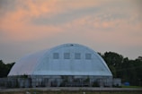 A large white structure with a rounded roof resembling a storage facility or warehouse. The building is situated in a rural setting with trees in the background and has a cloudy orange and blue sky overhead.