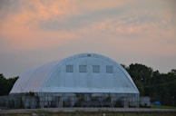 A large white structure with a rounded roof resembling a storage facility or warehouse. The building is situated in a rural setting with trees in the background and has a cloudy orange and blue sky overhead.