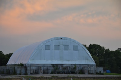 A large white structure with a rounded roof resembling a storage facility or warehouse. The building is situated in a rural setting with trees in the background and has a cloudy orange and blue sky overhead.