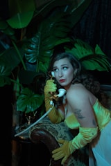 A woman wearing a vintage, glamorous yellow dress and gloves is using an old-fashioned telephone. She is positioned against a backdrop of large green leaves, creating a mysterious and retro atmosphere. The setting includes a patterned armrest, adding to the classic and elegant vibe.