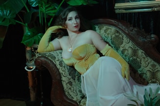 A woman is reclining on an ornate vintage sofa with a faint floral pattern. She wears a golden yellow corset and long matching gloves, paired with a sheer white skirt. Her hair is styled in soft curls, and she is surrounded by lush green plants, suggesting a tropical or elegant atmosphere.