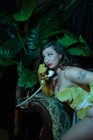 A woman in a vintage-style yellow outfit and long gloves is sitting on an ornately patterned armchair, holding a retro white rotary phone to her ear. Lush green leaves of large plants create a dramatic background, giving the scene an exotic vibe. Her expression is playful, with bright red lipstick complementing her look. A tattoo is visible on her shoulder.