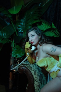 A woman in a vintage-style yellow outfit and long gloves is sitting on an ornately patterned armchair, holding a retro white rotary phone to her ear. Lush green leaves of large plants create a dramatic background, giving the scene an exotic vibe. Her expression is playful, with bright red lipstick complementing her look. A tattoo is visible on her shoulder.