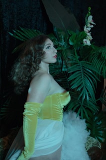 A woman with curly hair stands in front of lush green foliage, wearing a yellow velvet top and long gloves, with a sheer and flowing white garment. Her gaze is directed upwards, and the background features dark fabric and vibrant green leaves, along with light pink and white flowers.