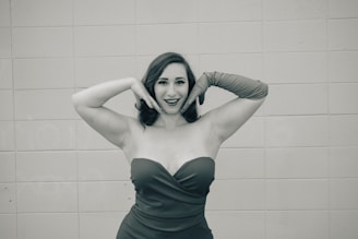A woman wearing a strapless dress and long gloves poses in front of a tiled wall with a playful expression, hands framing her face.