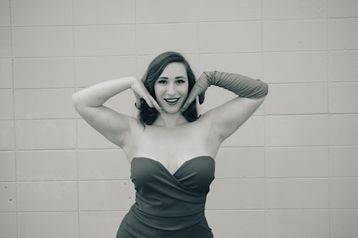 A woman wearing a strapless dress and long gloves poses in front of a tiled wall with a playful expression, hands framing her face.