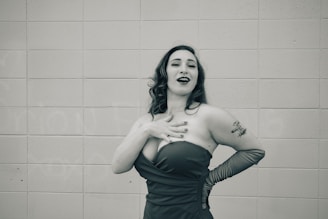 A woman stands in front of a tiled wall wearing an elegant strapless dress and long gloves. Her hair is styled in loose waves, and she has a tattoo on her upper arm. She appears to be laughing or singing, striking a confident pose with one hand on her chest and the other on her hip.