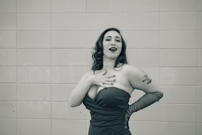 A woman stands in front of a tiled wall wearing an elegant strapless dress and long gloves. Her hair is styled in loose waves, and she has a tattoo on her upper arm. She appears to be laughing or singing, striking a confident pose with one hand on her chest and the other on her hip.