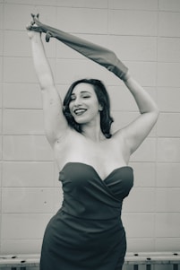 A woman in an elegant dress poses confidently with one arm raised, holding a long glove in the air. She has a joyful expression, with wavy hair and bold lipstick. The background features a simple tiled wall in a monochromatic color scheme.