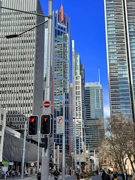 Tall modern skyscrapers with reflective glass facades dominate the urban landscape. Traffic lights and street signs are visible in the foreground, indicating a bustling city environment. Pedestrians walk along the street, and a tree with sparse foliage is on the side. A clear blue sky serves as the backdrop.