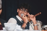 A person wearing glasses and a suit is speaking into a microphone while holding a gavel in one hand. Papers are scattered on the table in front of them, and a drum set is visible in the background.