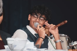 A person wearing glasses and a suit is speaking into a microphone while holding a gavel in one hand. Papers are scattered on the table in front of them, and a drum set is visible in the background.
