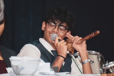 A person wearing glasses and a suit is speaking into a microphone while holding a gavel in one hand. Papers are scattered on the table in front of them, and a drum set is visible in the background.