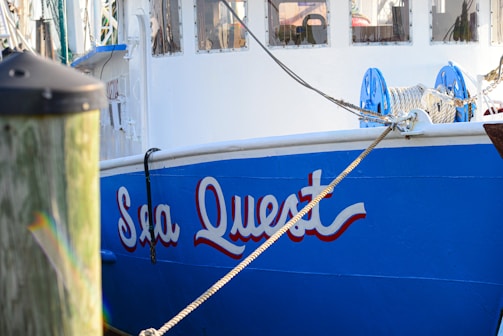 A docked boat with the name 'Sea Quest' painted in bold, blue lettering on the side. The boat features white and blue colors, with ropes and pulleys visible on the deck. Part of a wooden dock post is visible in the foreground.