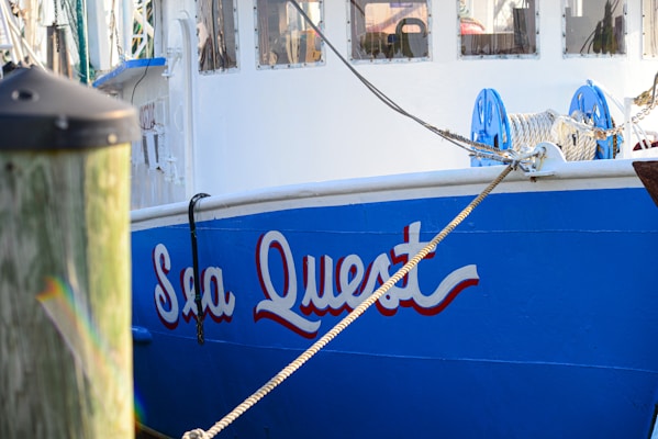 A docked boat with the name 'Sea Quest' painted in bold, blue lettering on the side. The boat features white and blue colors, with ropes and pulleys visible on the deck. Part of a wooden dock post is visible in the foreground.
