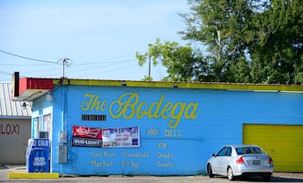 A small convenience store named 'The Bodega and Deli' with a blue exterior and yellow accents. It features signage advertising Bud Light and Budweiser beer promotions. Several advertisements and a Bud Light cooler are displayed outside the entrance. A silver car is parked in front of the store. The surrounding area includes green trees and a secondary building partially visible on the left.