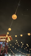 Lanterns line the night sky, creating a warm, inviting glow amidst the darkness. A red structure with decorative elements and signage is visible, suggesting a festive or cultural setting.