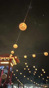 Lanterns line the night sky, creating a warm, inviting glow amidst the darkness. A red structure with decorative elements and signage is visible, suggesting a festive or cultural setting.