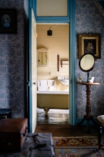 A vintage bathroom viewed through an open door, revealing a clawfoot bathtub with white towels hanging nearby. The room is decorated with patterned wallpaper and wooden accents, including a small table with a round mirror and a teacup. Framed pictures adorn the walls, while the floor is covered with a colorful rug.