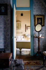 A vintage bathroom viewed through an open door, revealing a clawfoot bathtub with white towels hanging nearby. The room is decorated with patterned wallpaper and wooden accents, including a small table with a round mirror and a teacup. Framed pictures adorn the walls, while the floor is covered with a colorful rug.