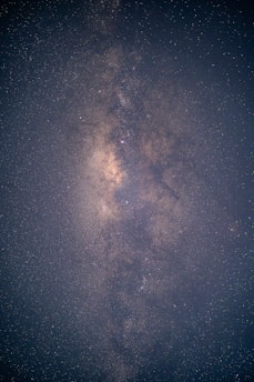 A striking image of the Milky Way galaxy stretching across the night sky. The dense star field features countless stars and clusters of varying brightness, creating a textured, glowing band. The background is a deep, dark blue, accentuating the luminous quality of the stars.