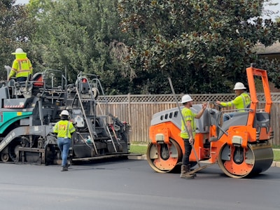 Several construction workers wearing safety vests and helmets are operating heavy machinery to lay asphalt on a road. One worker is driving an orange steamroller, while two others are managing a road paver machine. Trees and a wooden fence form the background.