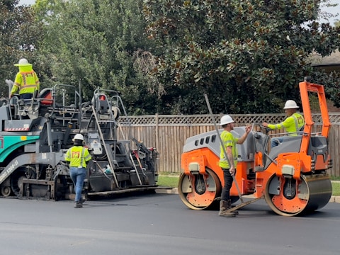 Several construction workers wearing safety vests and helmets are operating heavy machinery to lay asphalt on a road. One worker is driving an orange steamroller, while two others are managing a road paver machine. Trees and a wooden fence form the background.