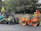 Several construction workers wearing safety vests and helmets are operating heavy machinery to lay asphalt on a road. One worker is driving an orange steamroller, while two others are managing a road paver machine. Trees and a wooden fence form the background.