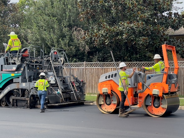 Several construction workers wearing safety vests and helmets are operating heavy machinery to lay asphalt on a road. One worker is driving an orange steamroller, while two others are managing a road paver machine. Trees and a wooden fence form the background.