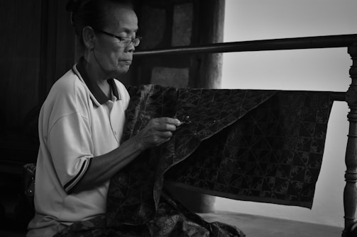 An elderly person with glasses is focused on meticulously crafting or sewing a piece of patterned fabric. The setting appears to be indoors with a railing and a wall in the background, creating a calm and intimate scene.