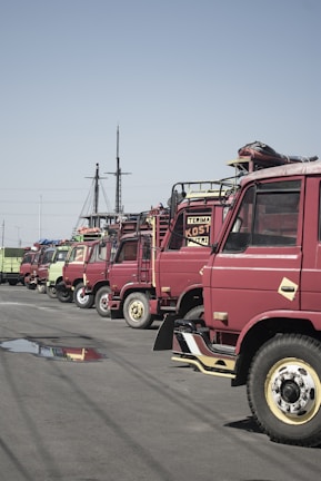 A row of parked red trucks is aligned along a street with a clear blue sky overhead. Some of the trucks have signs with text such as 'TERIMA' on them. There is a noticeable water puddle reflecting the trucks on the asphalt.