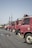 A row of parked red trucks is aligned along a street with a clear blue sky overhead. Some of the trucks have signs with text such as 'TERIMA' on them. There is a noticeable water puddle reflecting the trucks on the asphalt.