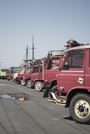 A row of parked red trucks is aligned along a street with a clear blue sky overhead. Some of the trucks have signs with text such as 'TERIMA' on them. There is a noticeable water puddle reflecting the trucks on the asphalt.