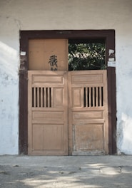 A pair of old wooden doors with a rustic appearance set into a wall. The wall is painted white and has some sign of wear and age. The doors are partially open, revealing greenery in the background. Above the doors, there is some writing or carvings, and one of the door panels has some black graffiti.