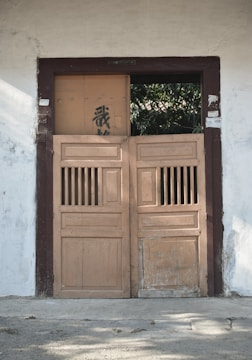 A pair of old wooden doors with a rustic appearance set into a wall. The wall is painted white and has some sign of wear and age. The doors are partially open, revealing greenery in the background. Above the doors, there is some writing or carvings, and one of the door panels has some black graffiti.