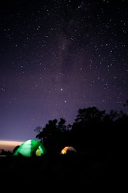 A starry night sky serves as a backdrop to a camping scene with two illuminated tents. The stars create a sparkling canvas above, while the tents emit a soft, warm glow in greens and yellows. Silhouettes of trees form a dark outline against the sky.