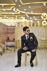 A well-dressed man sits on a golden chair in a lavish, illuminated setting. He is wearing a black suit with a patterned tie and an orange pocket square. He holds a phone in one hand and wears sunglasses and a large blue wristwatch. The background shows an opulently decorated interior with bright ceiling lights and ornate wall designs.