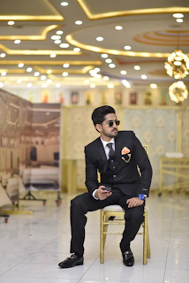 A well-dressed man sits on a golden chair in a lavish, illuminated setting. He is wearing a black suit with a patterned tie and an orange pocket square. He holds a phone in one hand and wears sunglasses and a large blue wristwatch. The background shows an opulently decorated interior with bright ceiling lights and ornate wall designs.