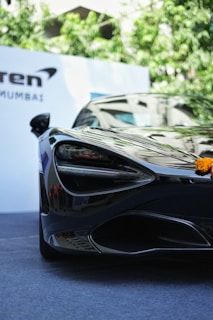 A sleek, black sports car with glossy finish and aerodynamic design is prominently displayed in an outdoor setting. The front of the car features sharp, angular headlights and a smooth, curved hood. There is a small marigold flower garland placed on the front of the vehicle. The background includes green foliage and a white banner with text.