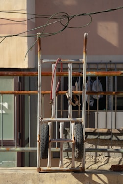 A metal hand truck with rubber tires is leaning against a railing. The frame shows signs of rust, and there are cables loosely draped over it. In the background, a beige wall and a window with bars and hanging clothes are visible.