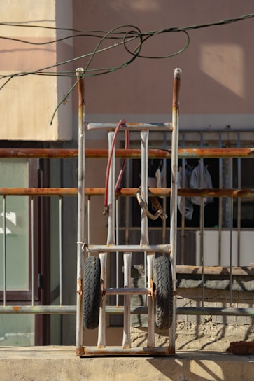 A metal hand truck with rubber tires is leaning against a railing. The frame shows signs of rust, and there are cables loosely draped over it. In the background, a beige wall and a window with bars and hanging clothes are visible.