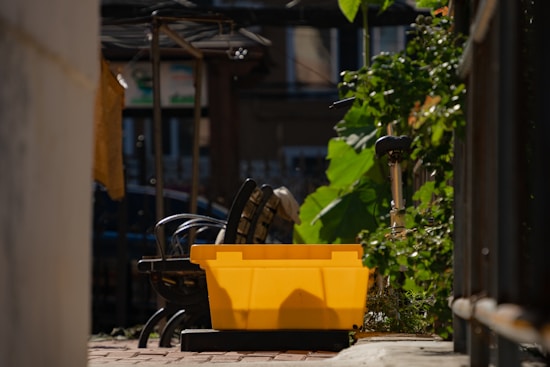 A bright yellow plastic box is placed on a pathway with a brick surface. To the left, metal chairs are stacked, and to the right, green leafy plants grow along a railing. In the background, parts of a bicycle and some buildings are visible.